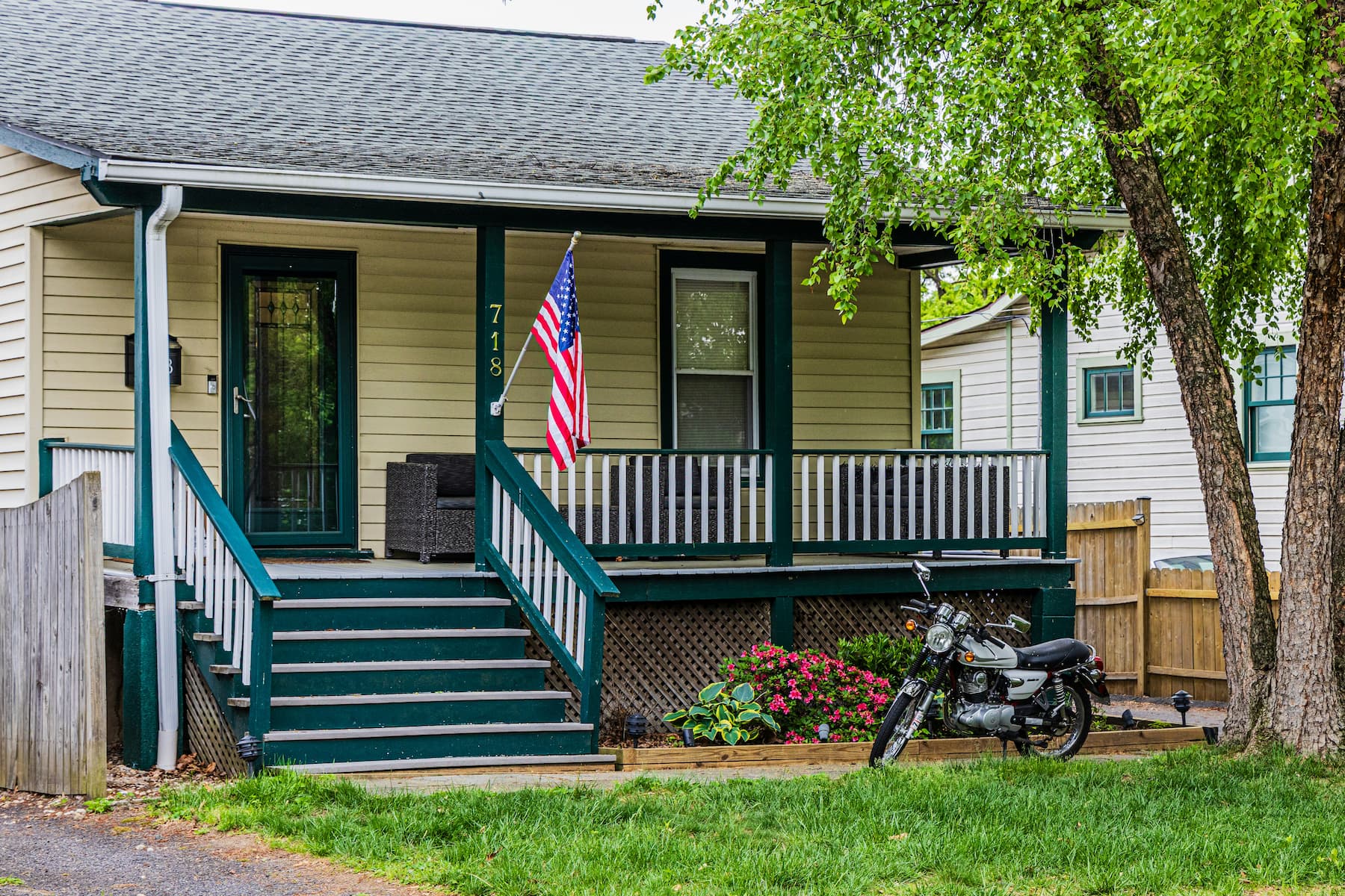 A modest American home with a front porch, lawn, and U.S. flag, with a vintage motorcycle parked out front.